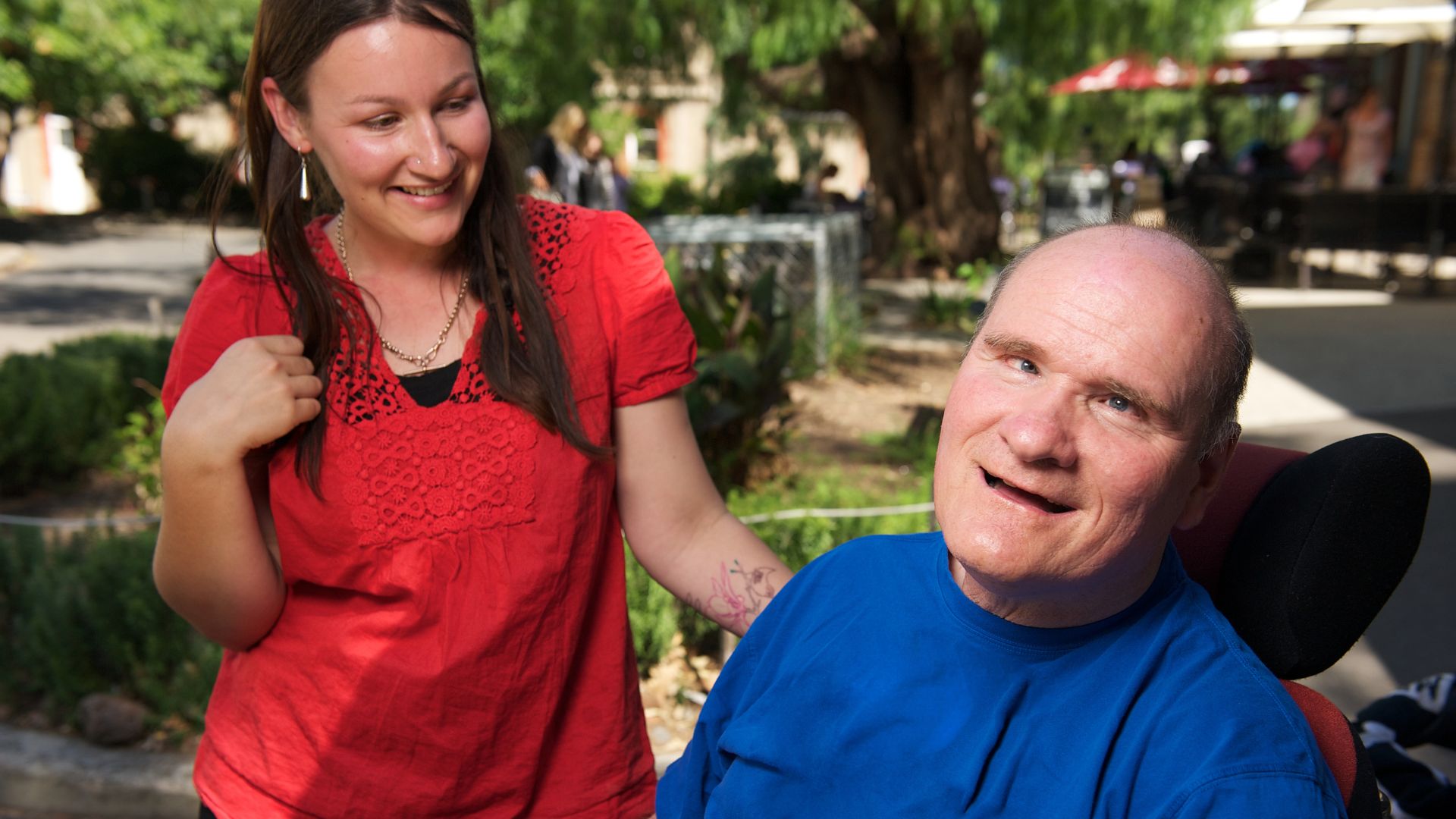 A lady in a red dress supporting a man in a wheelchair through independent living options.